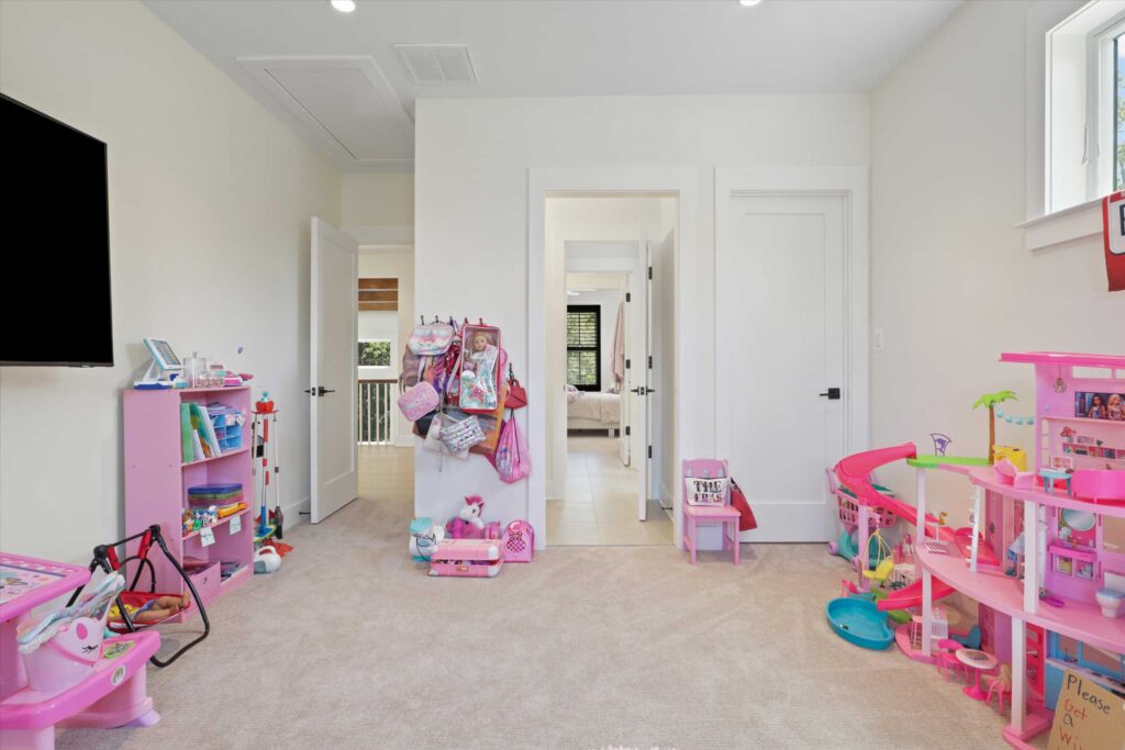 Bright carpeted bedroom utilized as a playroom, featuring pink toy storage, a dollhouse, and a wall-mounted television.