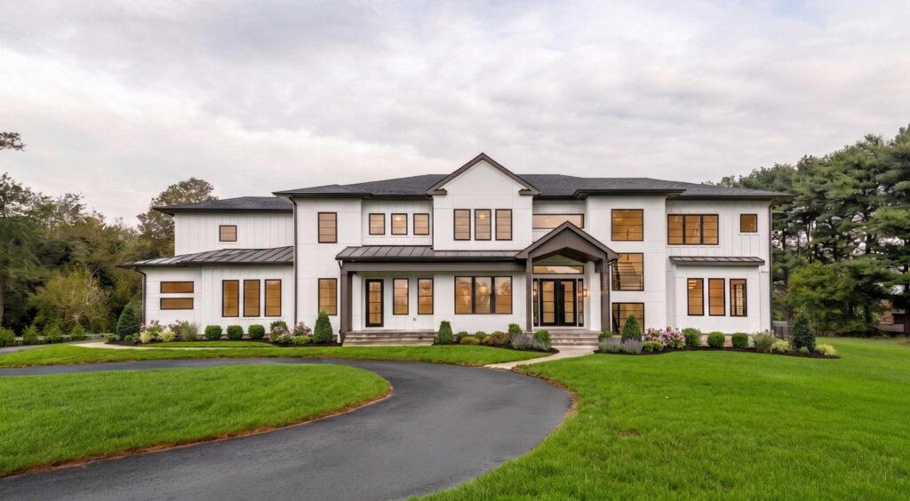 Front exterior view of a sprawling luxury modern farmhouse. The two-story residence features white vertical siding and flat panels with contrasting black window frames and a dark roof. A curved asphalt driveway leads through a green lawn to a central covered entrance with a gabled portico and stone foundation skirting.
