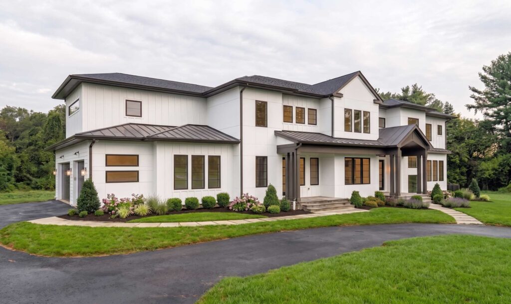 Angled front exterior view of a sprawling luxury modern farmhouse. The two-story residence features white vertical siding and flat panels with contrasting black window frames and a dark roof. A curved asphalt driveway leads through a green lawn to a central covered entrance with a gabled portico and stone foundation skirting.