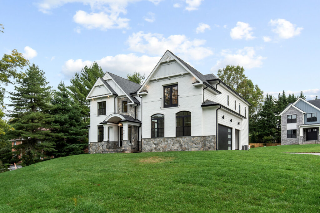Exterior view of a white brick luxury home featuring black window frames, stone foundation accents, and a side-loading garage situated on a grassy hill.