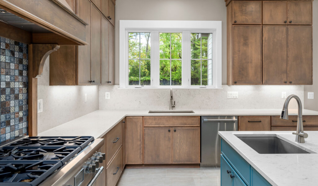 Close-up of a kitchen corner featuring a stainless steel gas cooktop, a blue and brown mosaic tile backsplash, and a custom wood range hood.