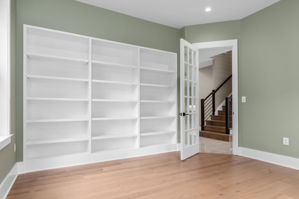 Home office or study with sage green walls, floor-to-ceiling built-in white bookshelves, hardwood flooring, and a glass-paned door.