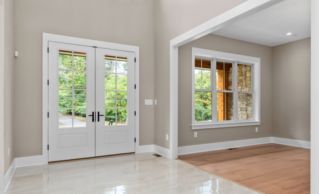 Bright foyer entrance with glossy tile flooring, double white glass-paneled front doors, and a wide opening leading to a room with hardwood floors.