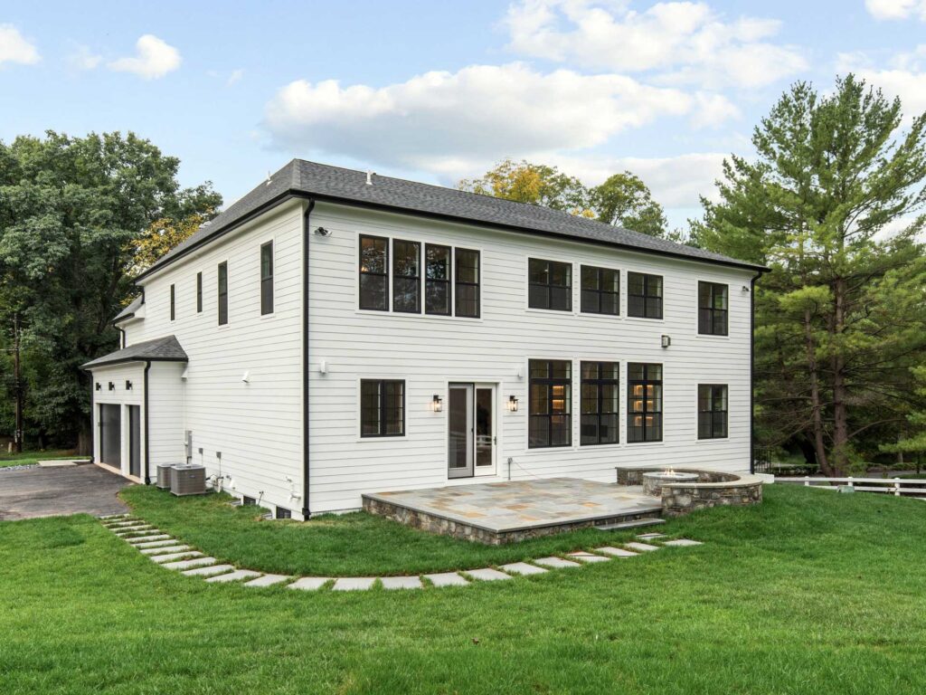 Rear exterior view of a white modern farmhouse showing a stone patio, fire pit, and large black-framed windows overlooking the lawn.