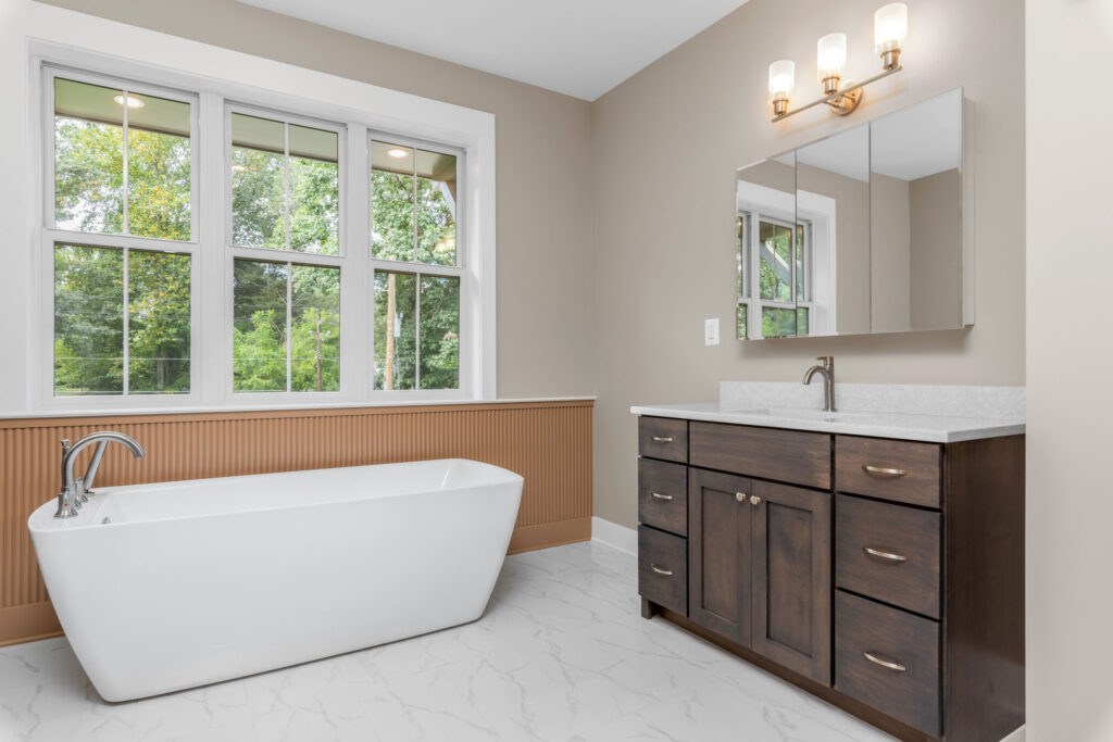 Bathroom vanity area featuring dark wood cabinets and a triple mirror, adjacent to a freestanding white bathtub against a wainscoted wall.