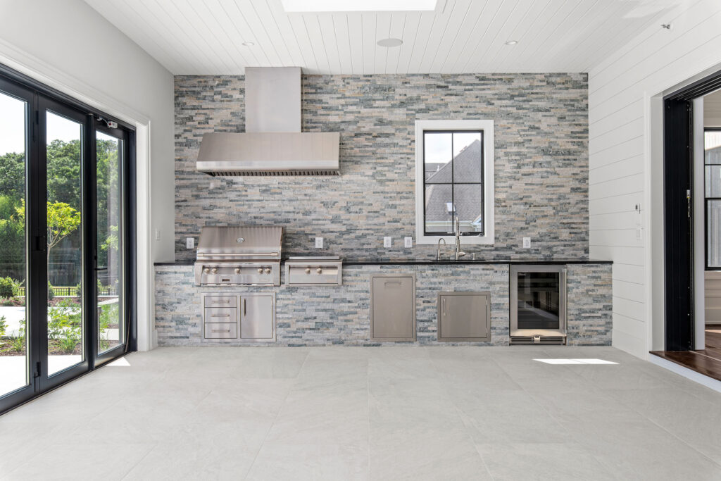 a sophisticated indoor-outdoor kitchen/pool house cabana designed for entertaining. The focal point is a floor-to-ceiling stacked stone feature wall that houses a built-in professional-grade stainless steel grill with a large range hood, a prep sink, and a stainless steel beverage refrigerator. The room features a white shiplap ceiling with recessed lighting and skylights, light gray large-format tile flooring, and a wall of black-framed accordion glass doors that fold open to seamlessly connect the space with the outdoor patio and pool area.