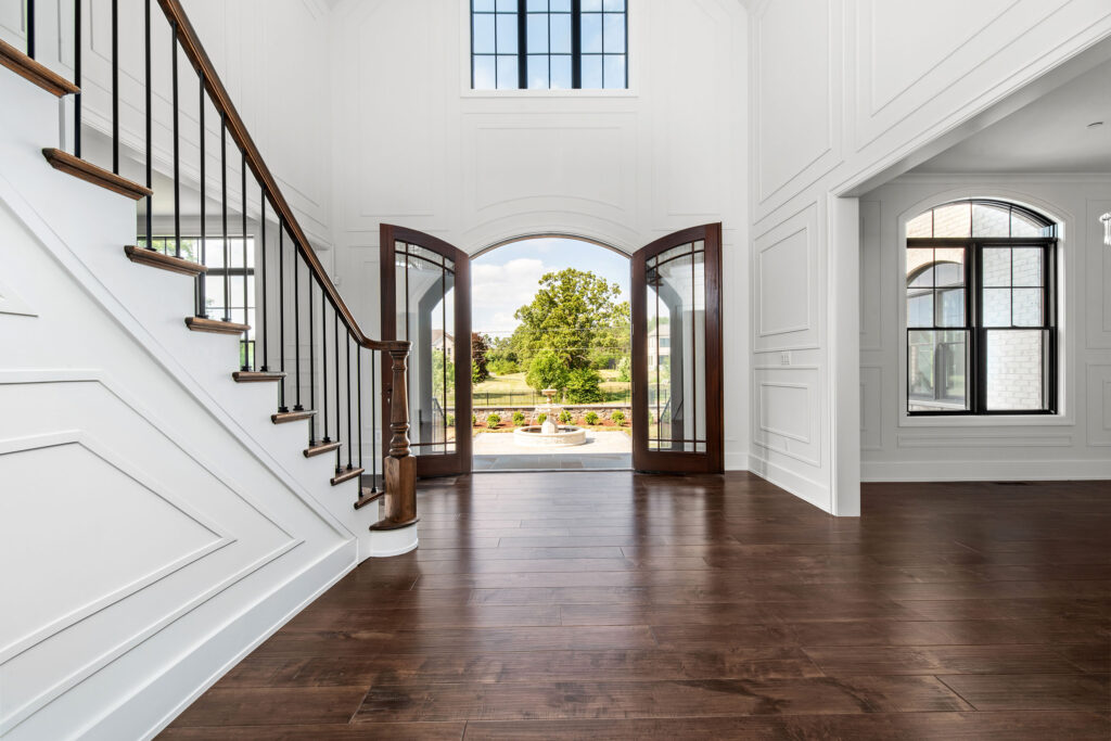 Interior view of a grand foyer looking outward through open double arched wood doors that frame a direct view of the exterior tiered stone fountain and front yard. The entry features dark wide-plank hardwood floors and towering white-paneled walls. To the left, the staircase is visible with dark wood treads and white risers, while a black-framed arched window accents the wall on the right.