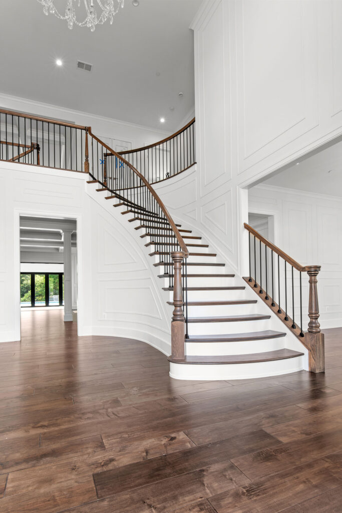 Interior view of a grand two-story foyer featuring a sweeping curved staircase with dark wood treads, white risers, and black iron balusters. The space is defined by extensive white wall paneling, wide-plank hardwood flooring, and a crystal chandelier. An open doorway to the left provides a sightline through to the rear entertainment wing and the outdoor pool area beyond.