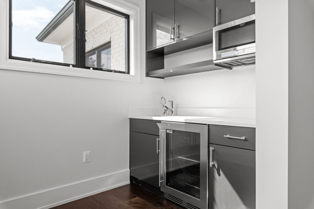 Close-up interior view of a sleek butler's pantry wet bar featuring high-gloss dark gray cabinetry and white quartz countertops. The space is equipped with a stainless steel prep sink, a built-in glass-door beverage refrigerator, and a microwave niche. Natural light enters through a black-framed window that overlooks the home's white brick exterior, while dark hardwood flooring completes the modern design.
