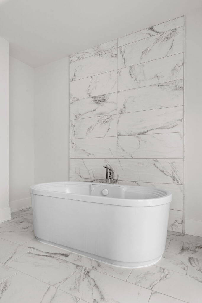Close-up interior view of a modern white freestanding bathtub positioned against a striking floor-to-ceiling accent wall featuring large-format white marble-look tiles. The tub includes a sleek chrome faucet, and the surrounding floor is finished with matching marble-style tiling, creating a seamless, spa-like aesthetic.