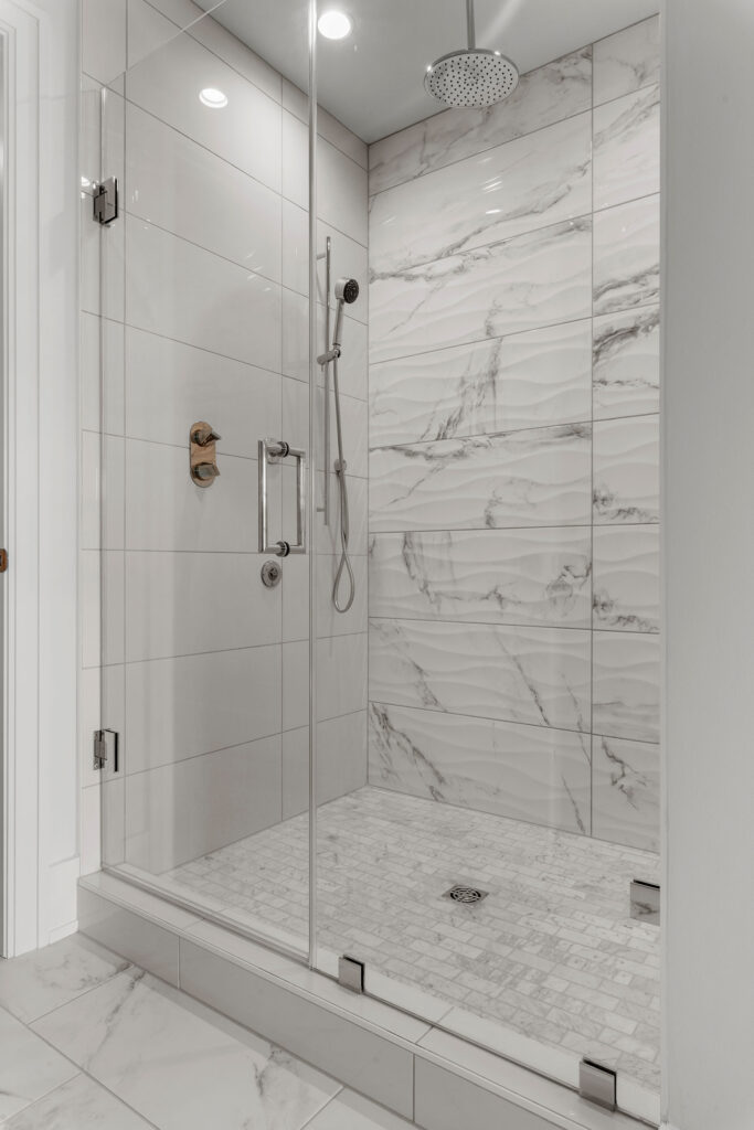 Interior view of a luxurious walk-in shower enclosed by a frameless glass door with chrome hardware. The shower features large-format white marble-look wall tiles and a matching mosaic tile floor. It is equipped with modern chrome fixtures, including a ceiling-mounted overhead rainfall showerhead and a separate handheld sprayer on a slide bar.