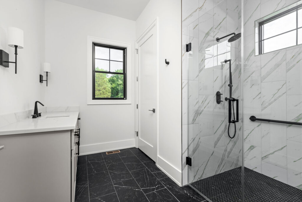 Interior view of a stylish bathroom featuring a light gray vanity with a white countertop and matte black faucet. The room is grounded by dark charcoal marble-look floor tiles and includes a walk-in glass shower with white marble-style wall tiles, a black mosaic floor, and matte black plumbing fixtures. A single black-framed window provides natural light and looks out onto green trees.