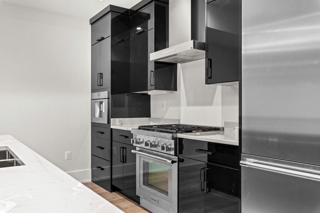 Close-up interior view of a modern kitchenette featuring high-gloss black cabinetry with sleek black hardware. The space is equipped with professional-grade stainless steel appliances, including a gas range oven, a ventilation hood, and a refrigerator. White marble-look quartz countertops and light wood-style flooring complete the contemporary design.