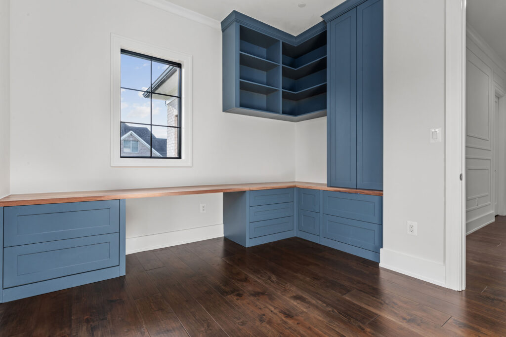 custom slate blue built-in cabinetry and a natural wood butcher block desktop. The workspace includes ample storage with drawers, corner open shelving, and a tall utility cabinet. Dark hardwood flooring contrasts with the crisp white walls, while a black-framed window allows natural light into the room.