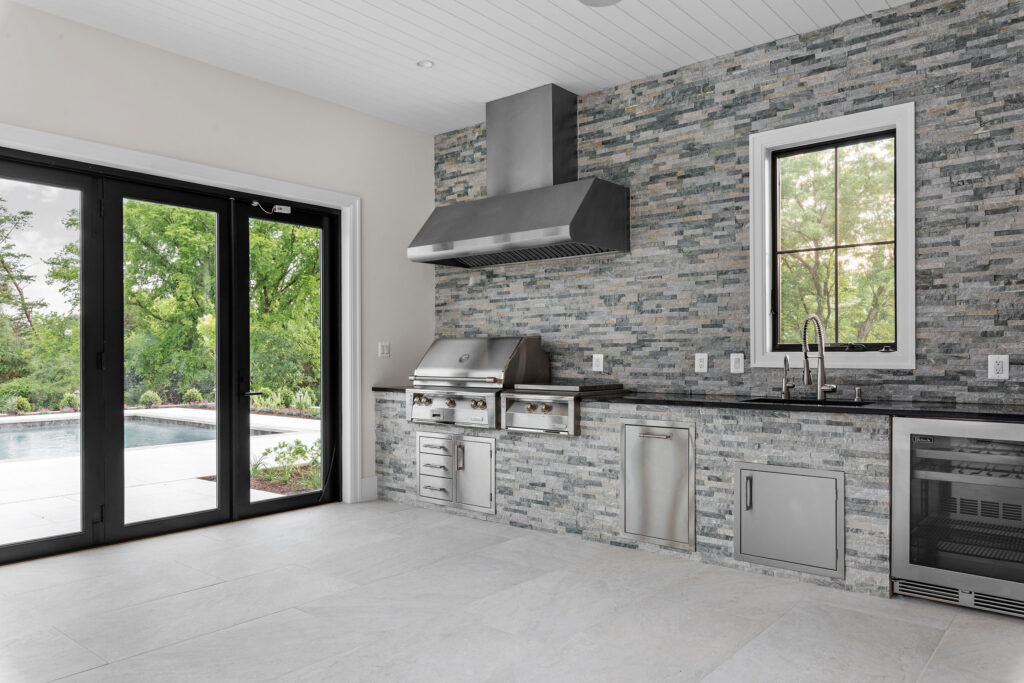 Interior view of a luxury indoor-outdoor kitchen featuring a stacked stone wall with a built-in stainless steel grill, range hood, and beverage refrigerator. Large black-framed folding glass doors on the left open to the swimming pool patio, creating a seamless transition to the outdoors. The space is finished with light gray tile flooring, a white shiplap ceiling, and a black countertop with a prep sink.