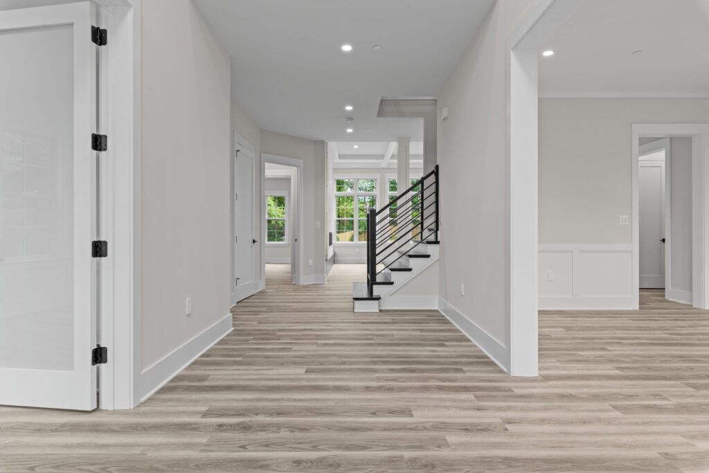 Bright open hallway with light wood flooring, white wainscoting, and a staircase featuring modern horizontal black metal railings.