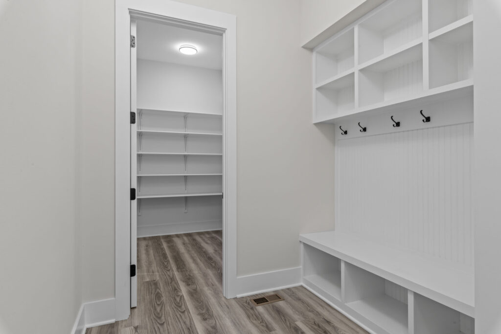 Mudroom area featuring white built-in storage lockers with coat hooks and cubbies, adjacent to a walk-in pantry with open shelving.