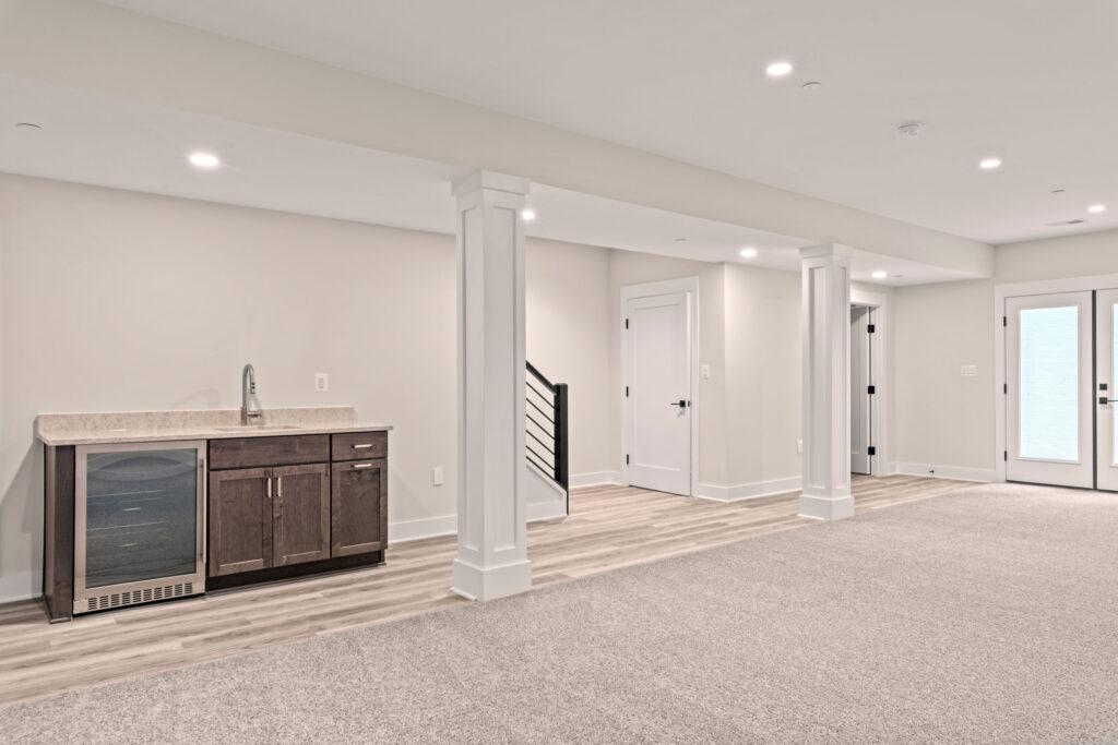 Spacious finished basement featuring a wet bar with dark cabinetry and a wine fridge, light grey carpeting, and double doors leading outside.