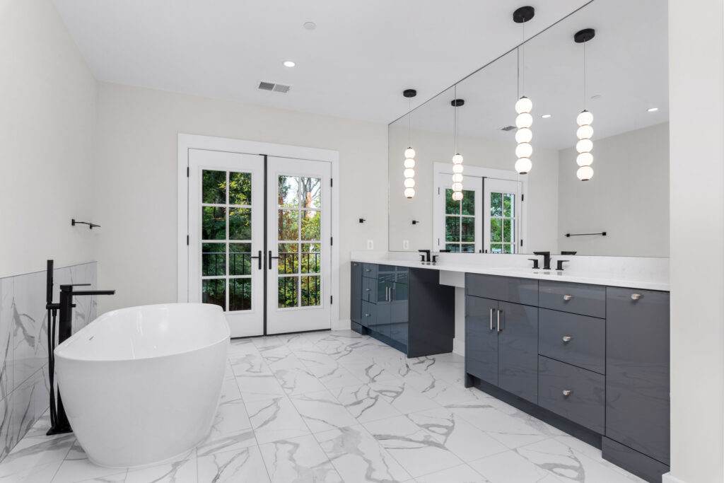 Spa-like bathroom interior with a freestanding white bathtub, dark grey glossy vanity, marble-look tile flooring, and double doors leading to a balcony.