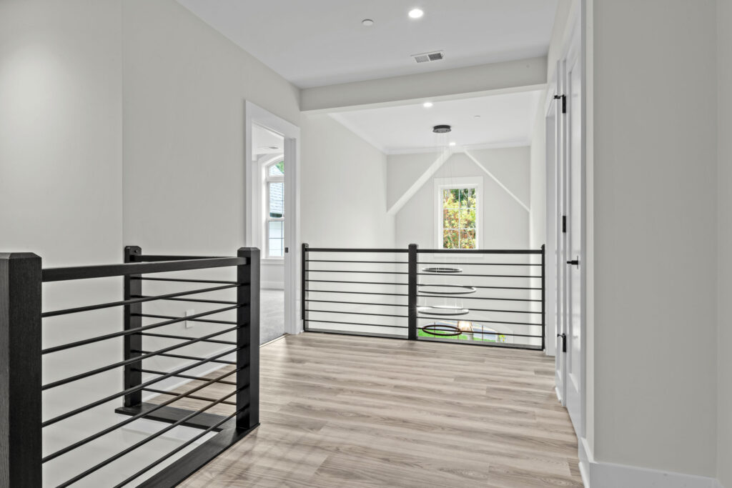 Upper-level hallway landing with black metal railings, looking out toward a window and the foyer chandelier.