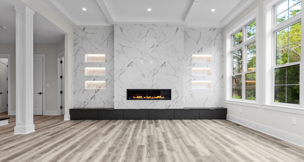 Empty great room featuring a floor-to-ceiling marble-look tile fireplace wall with illuminated display niches and large windows.