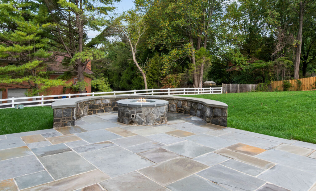 Outdoor stone patio featuring a built-in circular fire pit with a curved stone seating wall, set against a backdrop of green grass and trees.