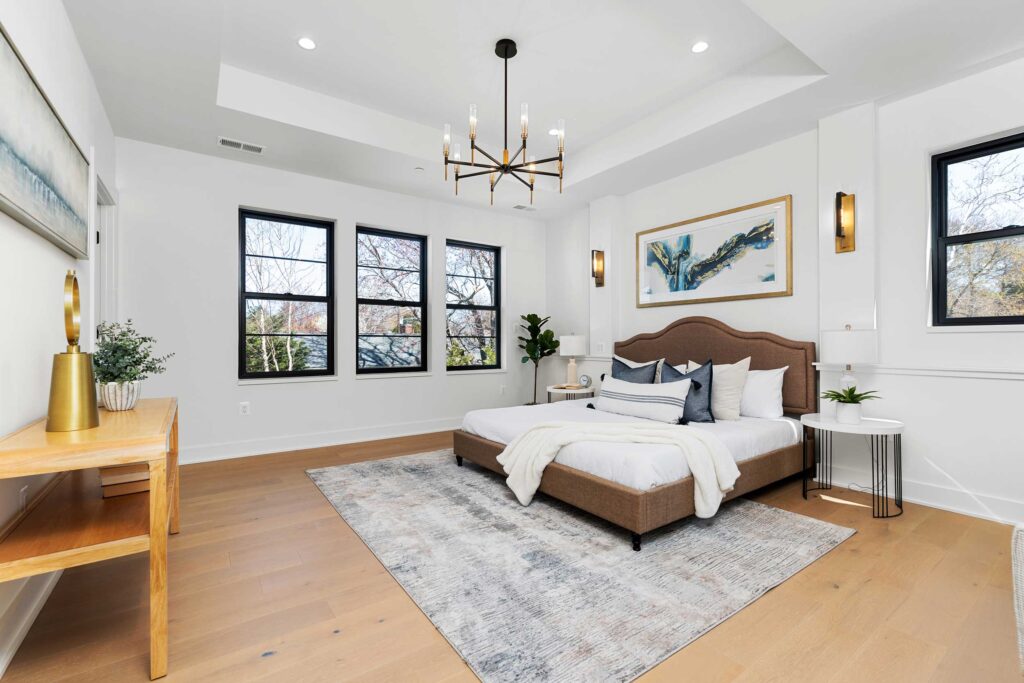 Primary bedroom featuring a brown upholstered bed with blue accent pillows, flanked by three large black-framed windows, brass wall sconces, and a contemporary fiddle leaf fig plant.