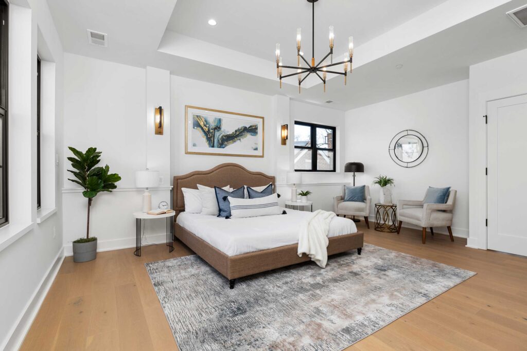 Wide view of a bright primary bedroom suite showing the layout of the bed, a seating area with two armchairs and a round mirror, and the intricate tray ceiling detail.