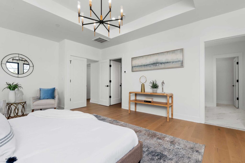 Spacious primary bedroom with a tray ceiling, black chandelier, light hardwood floors, and a seating nook with a grey armchair.