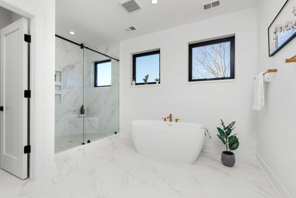 Wide view of a primary bathroom suite featuring a glass walk-in shower, a freestanding tub under a window, and light marble-patterned tile flooring.