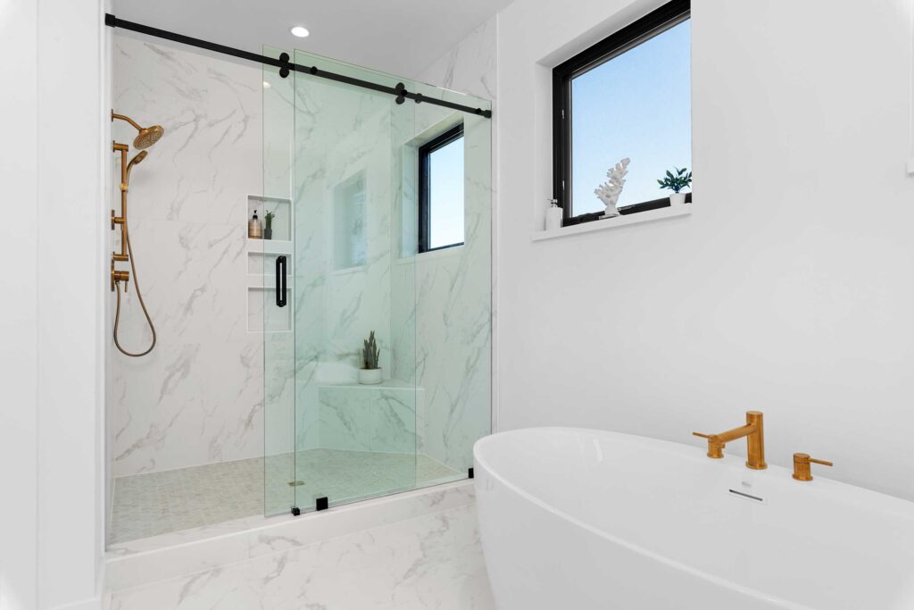 Bright bathroom corner showcasing a white freestanding soaking tub with gold fixtures beside a glass-enclosed walk-in shower with marble-look tile and black hardware.