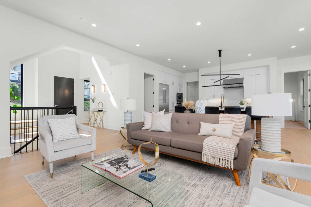 Open-concept living area with light hardwood floors, a mid-century modern grey sofa, and clear sightlines into the kitchen and stairwell.