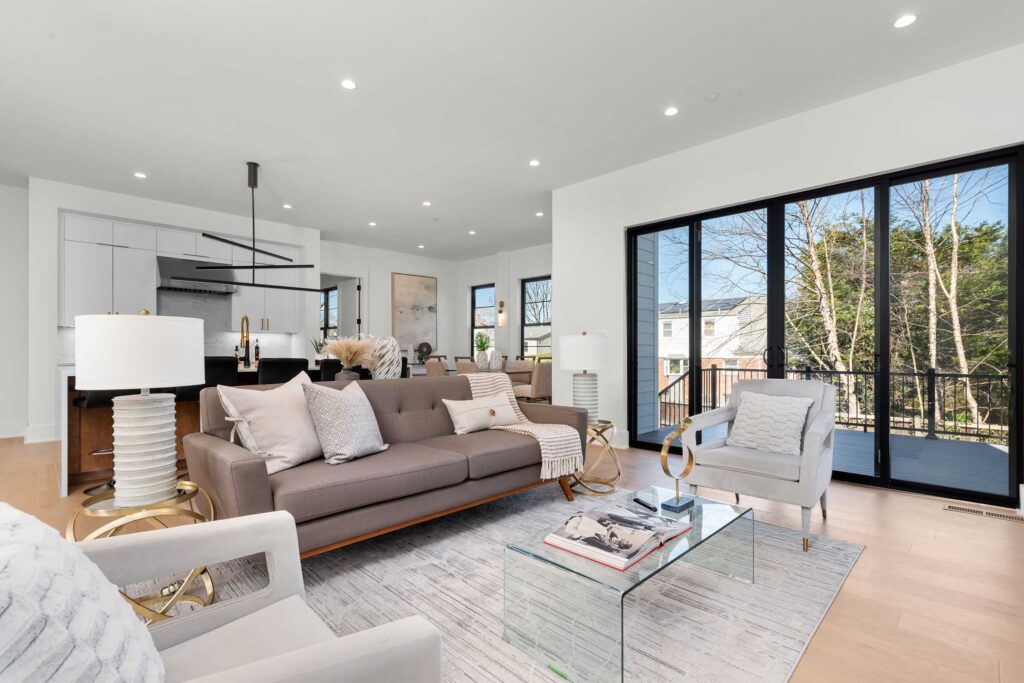 Sunlit living room interior with a grey sofa and velvet armchair facing a wall of floor-to-ceiling sliding glass doors that open onto a backyard deck.
