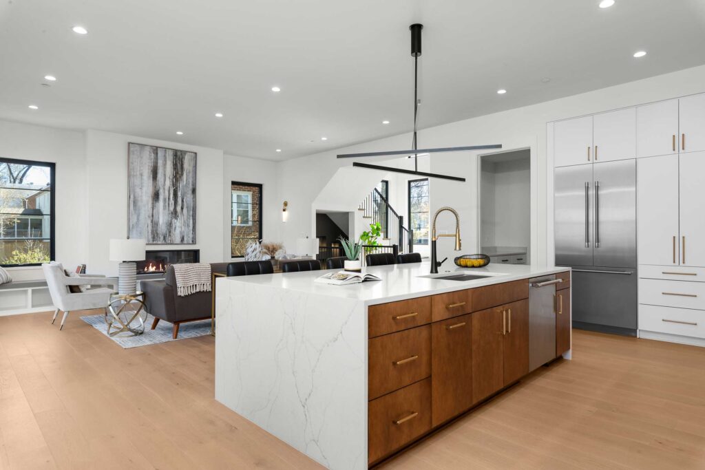 View from the kitchen island showing a gold gooseneck faucet and deep sink, looking out into an open-concept living room with a modern gas fireplace.
