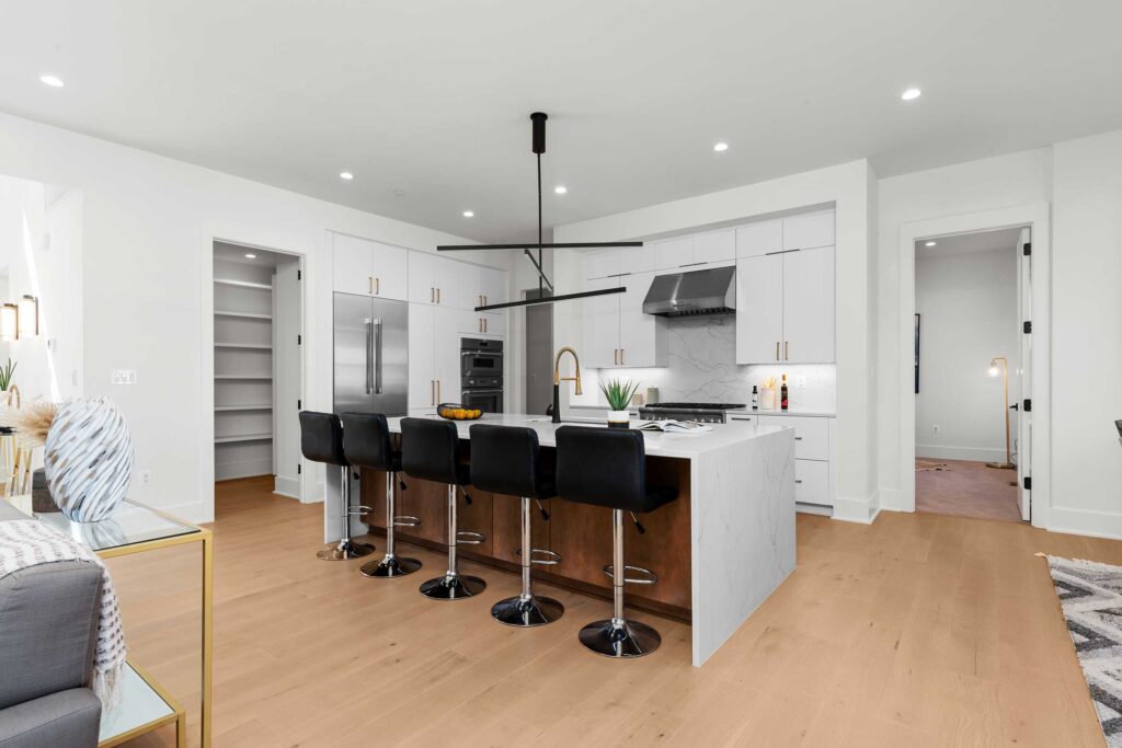 Full view of a modern kitchen layout including a large central island, stainless steel refrigerator, and a glimpse into a walk-in pantry with white shelving.