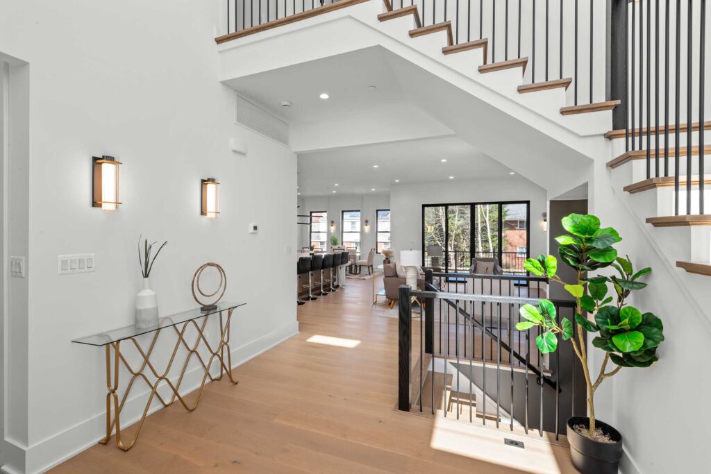 View from the hallway showing a modern staircase with open wood treads and black railing, leading down to a bright open-concept living and kitchen area.