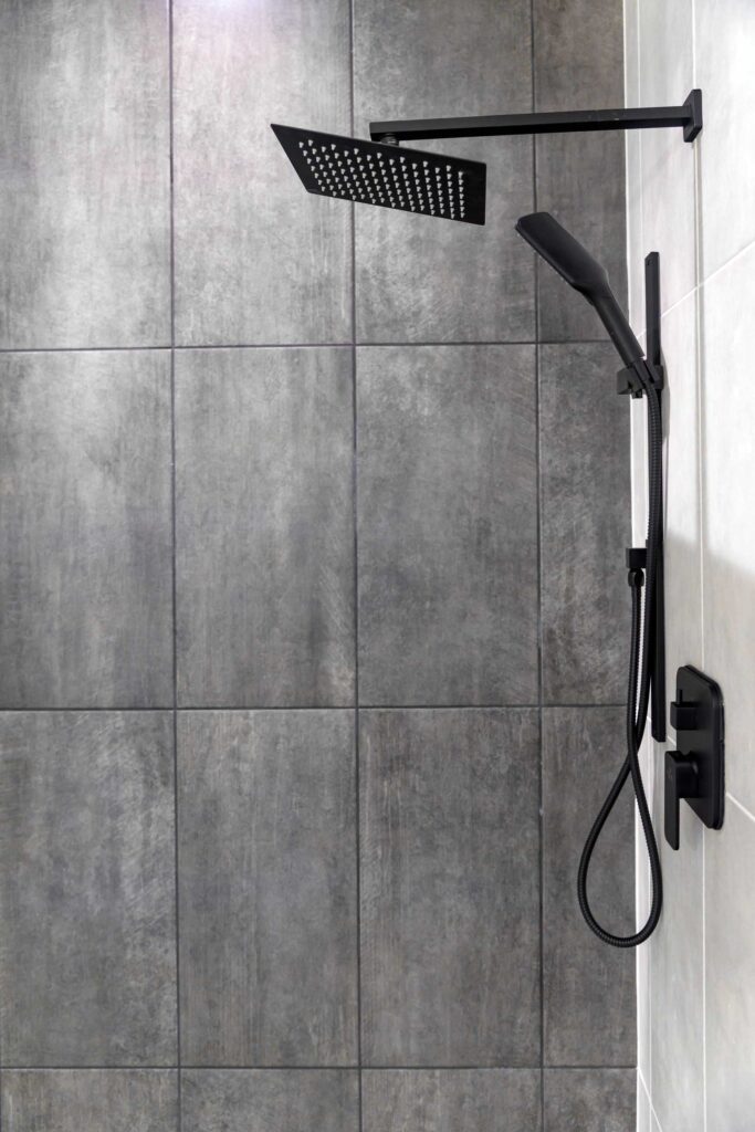 A modern shower system featuring matte black fixtures, including a square overhead rain shower head and a handheld wand on a vertical slide bar. The fixtures are mounted against a wall of large, textured gray tiles which meets a white tiled wall on the right.