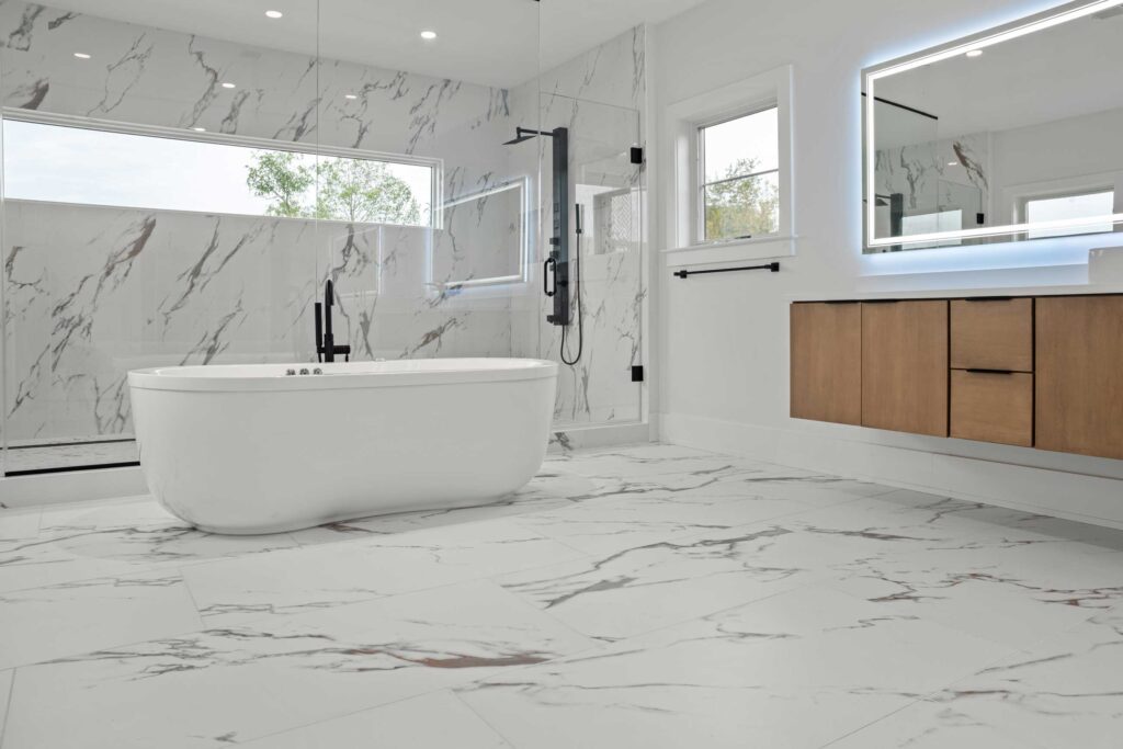 Angled view of a spacious luxury bathroom featuring large-format white marble tile flooring with grey veining. A curved freestanding soaking tub sits in front of a glass-enclosed walk-in shower equipped with matte black fixtures and a transom window. On the right, a floating light wood vanity is mounted below a large backlit mirror, accented by black hardware.