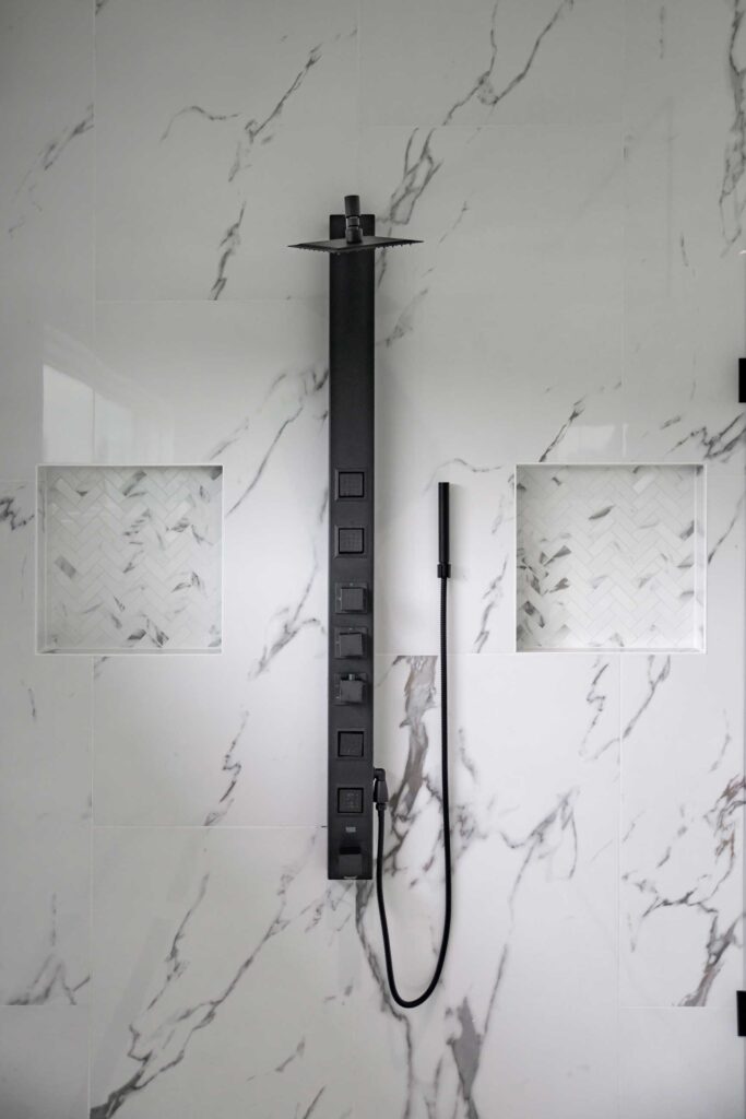 Close-up detail of a modern matte black shower panel system featuring a rain shower head, multiple body jets, and a handheld sprayer wand. The fixture is mounted on a white marble-look tile wall and is flanked by two symmetrical recessed shower niches with herringbone mosaic tile backing.