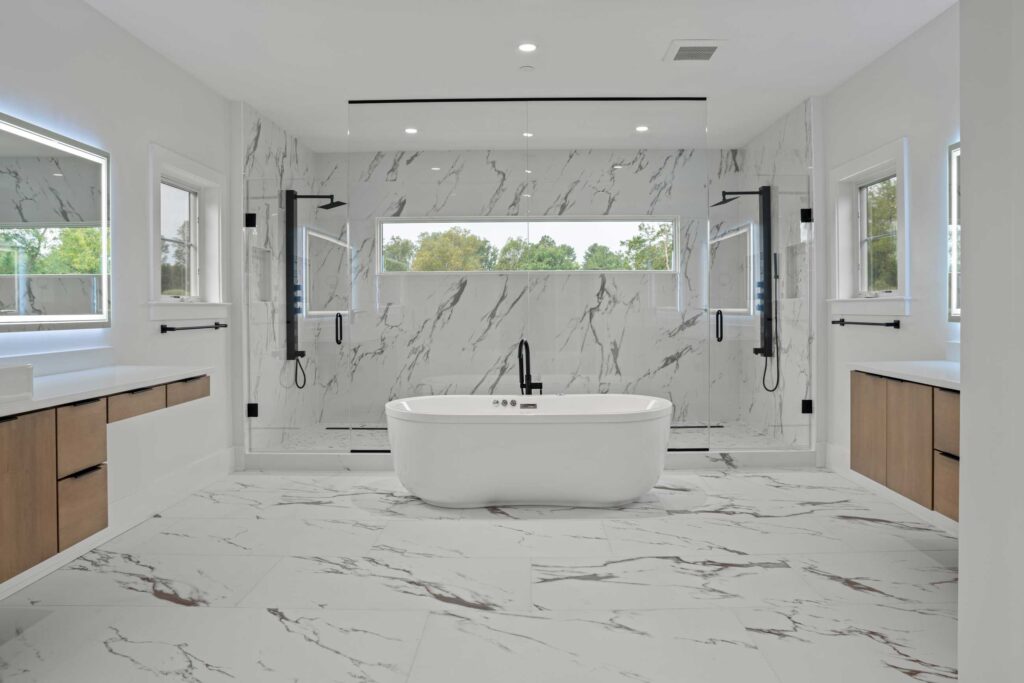 A spacious, modern luxury bathroom featuring white marble flooring and walls with grey veining. A white freestanding soaking tub sits centrally in front of a large, glass-enclosed walk-in shower with dual matte black fixtures. Symmetrical floating wood vanities with backlit mirrors line the side walls.