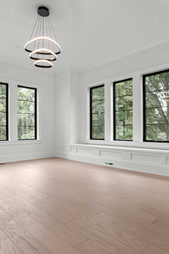 A spacious, bright bedroom featuring light hardwood flooring and crisp white walls. Large black-framed windows on adjacent walls flood the room with natural light and frame views of mature trees. A built-in white window seat runs the length of the triple window set on the right wall, while a modern, three-tiered black ring chandelier hangs from the ceiling.