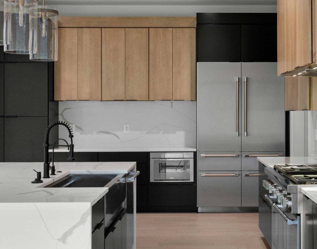 Modern kitchen featuring a large white quartz waterfall island with a matte black commercial-style faucet. The design showcases two-tone cabinetry with light wood uppers and flat-panel black lowers, a matching full-height slab backsplash, and high-end stainless steel appliances including a built-in refrigerator.