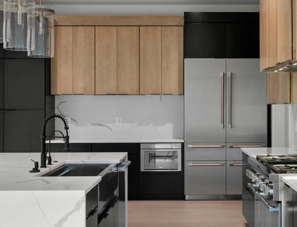 Modern kitchen featuring a large white quartz waterfall island with a matte black commercial-style faucet. The design showcases two-tone cabinetry with light wood uppers and flat-panel black lowers, a matching full-height slab backsplash, and high-end stainless steel appliances including a built-in refrigerator.