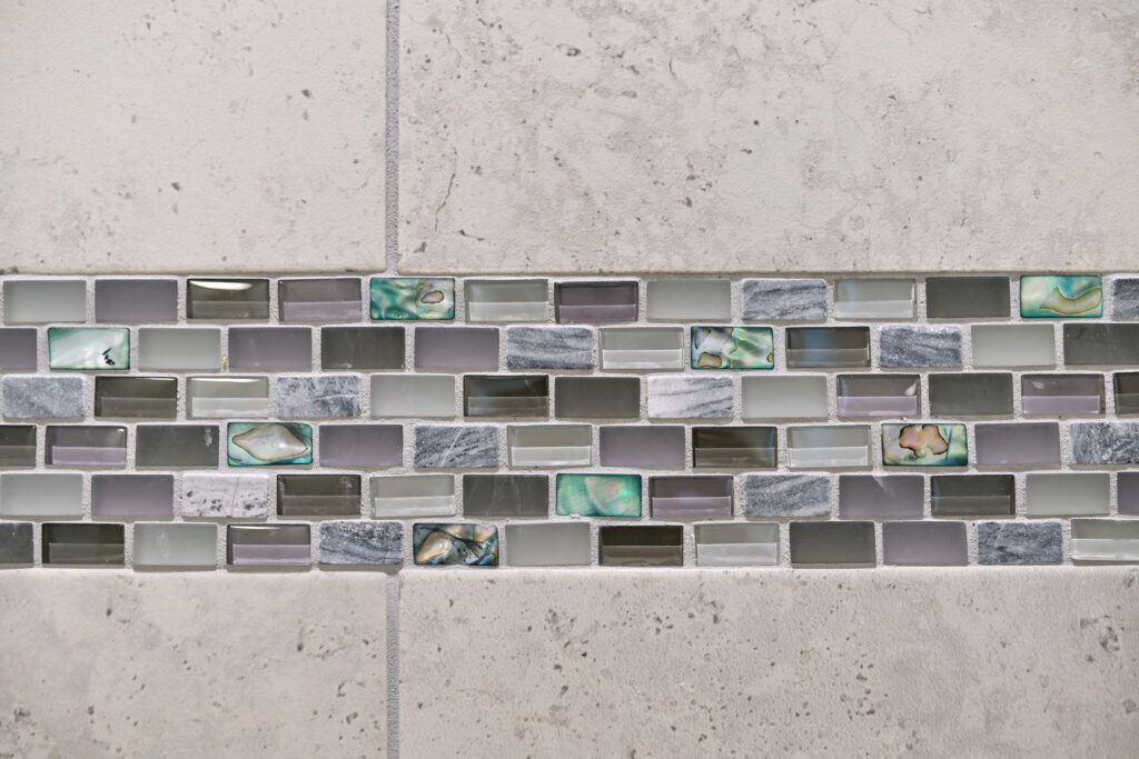 Close-up detail of beige stone bathroom tile featuring a decorative horizontal mosaic accent band with glass and abalone shell tiles.