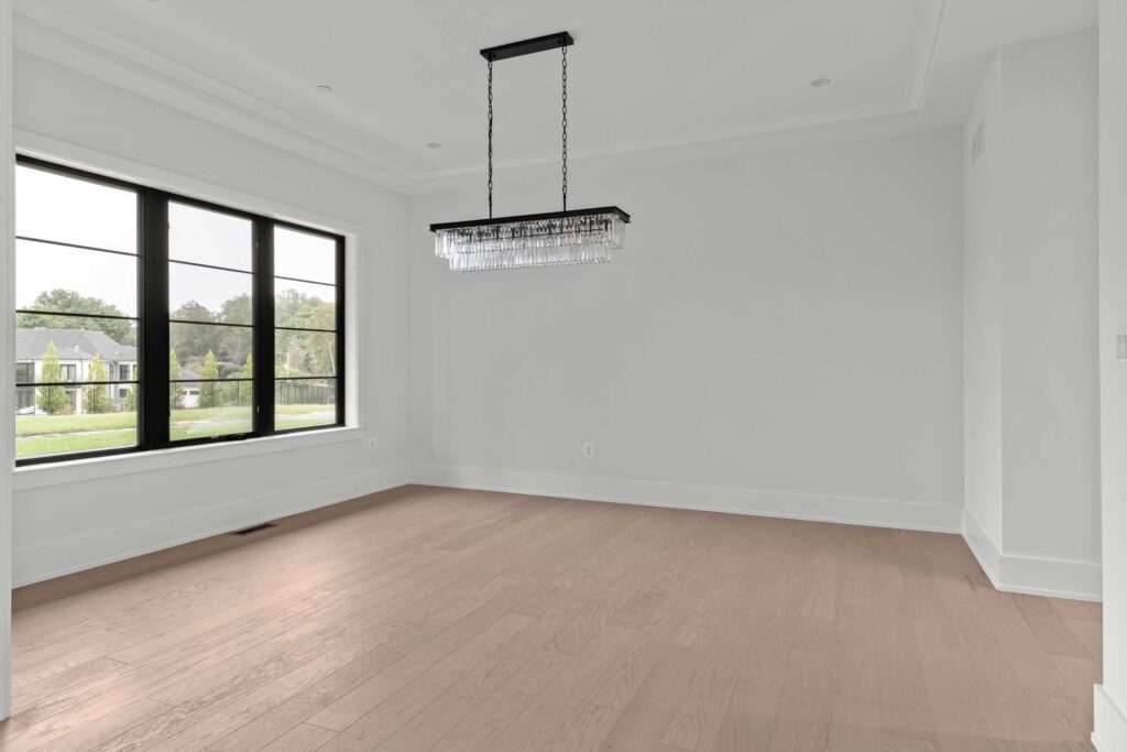 A bright, formal dining room featuring light hardwood flooring and clean white walls. A rectangular, modern crystal chandelier with black hardware hangs from the ceiling. To the left, a large black-framed window provides natural light and a view of the front lawn and neighboring homes.