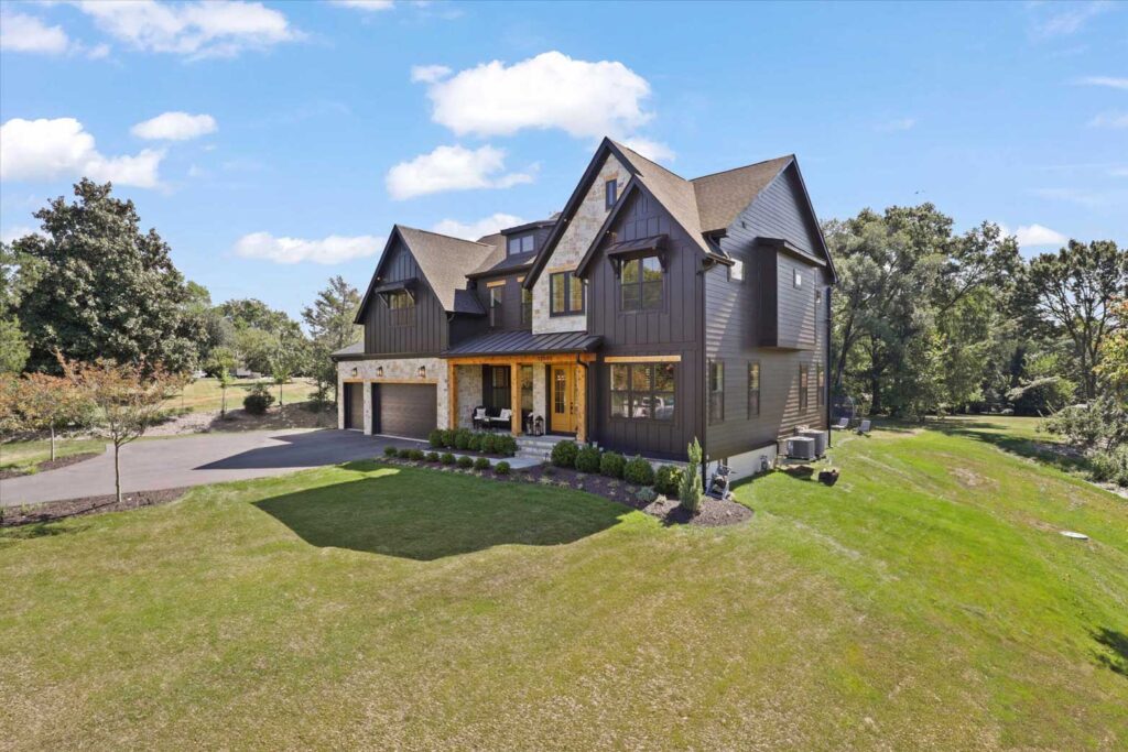 Angled exterior view of a custom home showcasing dark siding, stone veneer accents, a covered front porch with timber columns, and a three-car garage.