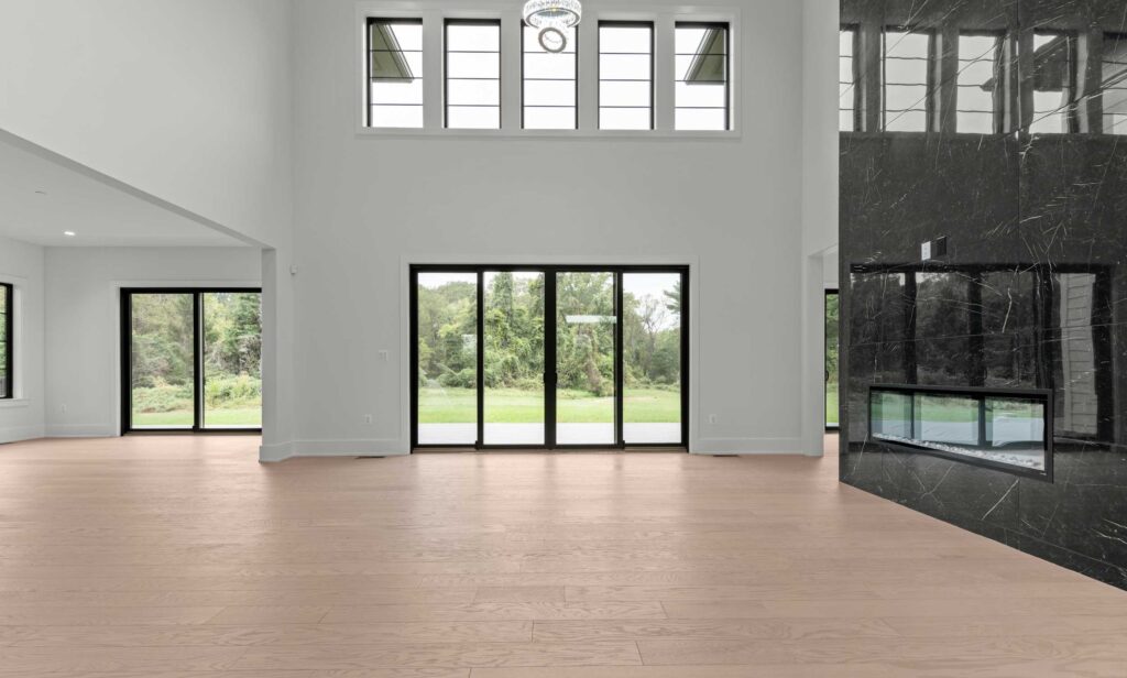A grand, double-height family room featuring light hardwood flooring and crisp white walls. To the right, a massive floor-to-ceiling black marble feature wall houses a sleek, linear gas fireplace. The rear wall showcases large black-framed sliding glass doors topped by a row of four clerestory windows, flooding the space with natural light and framing views of the green backyard.