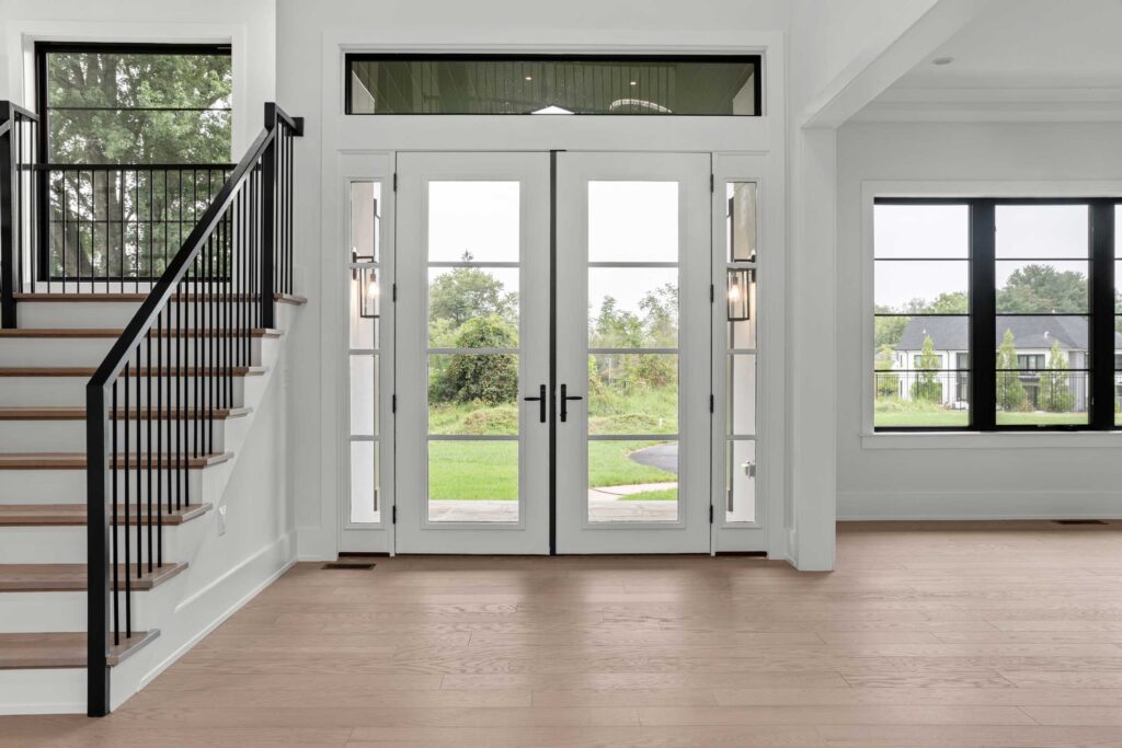 A spacious, modern foyer featuring light hardwood floors and crisp white walls. Centered are white double entry doors with glass panes and black hardware, flanked by sidelights and a transom window. To the left, a staircase ascends with wood treads and a black metal railing, while a large black-framed window on the right overlooks a green lawn and neighboring houses.