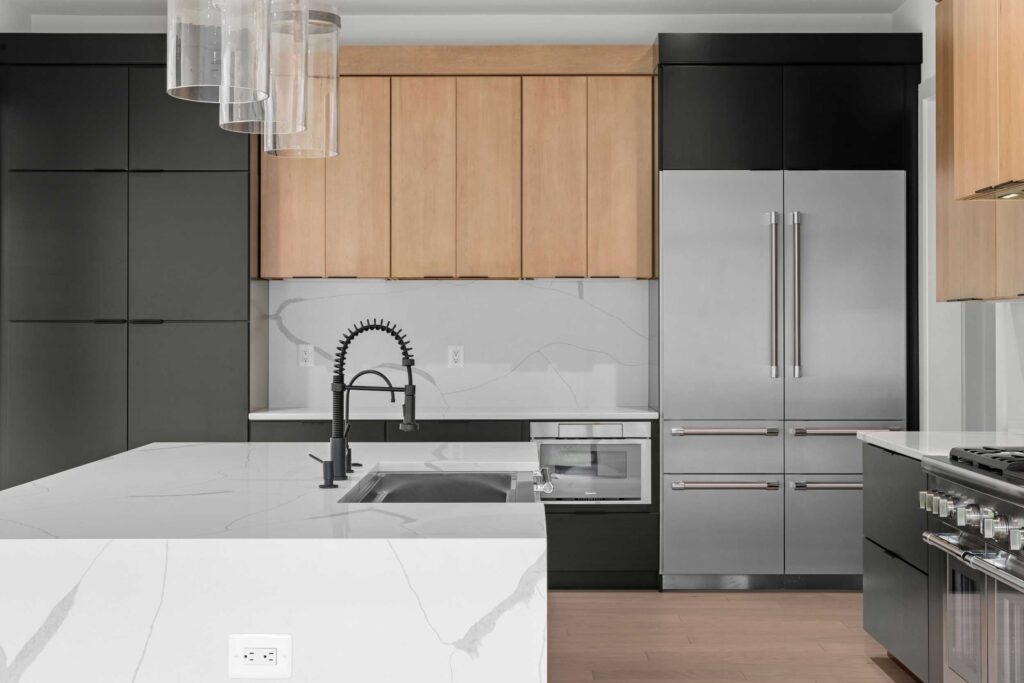 Modern kitchen featuring a large white quartz waterfall island with a matte black commercial-style faucet. The design showcases two-tone cabinetry with light wood uppers and flat-panel black lowers, a matching full-height slab backsplash, and high-end stainless steel appliances including a built-in refrigerator.