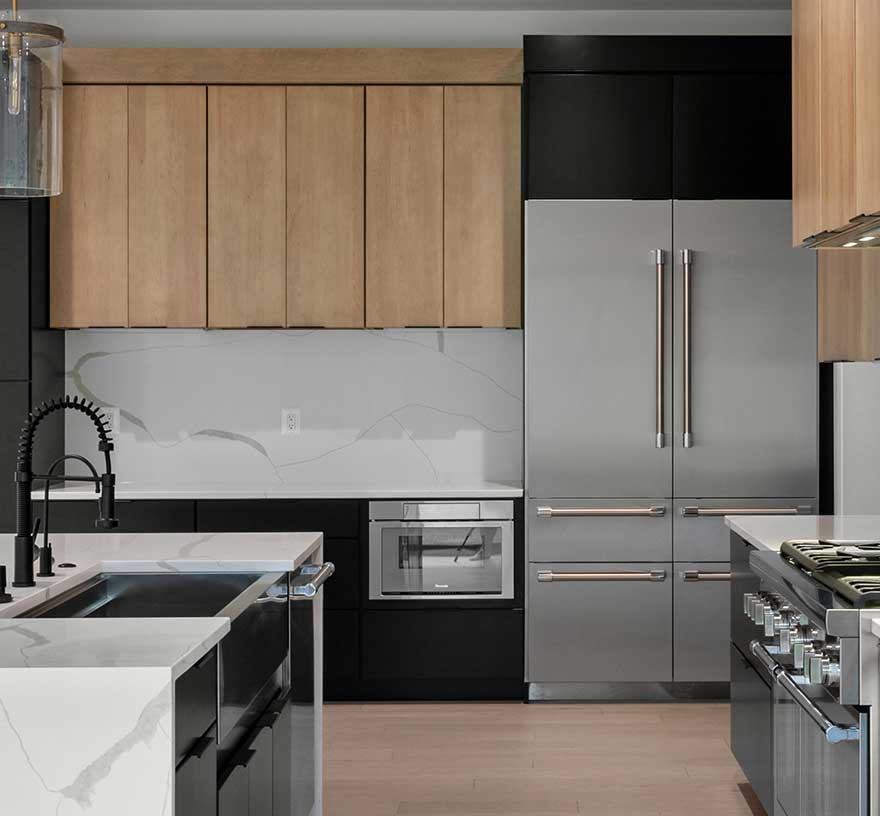 Modern kitchen featuring a large white quartz waterfall island with a matte black commercial-style faucet. The design showcases two-tone cabinetry with light wood uppers and flat-panel black lowers, a matching full-height slab backsplash, and high-end stainless steel appliances including a built-in refrigerator.